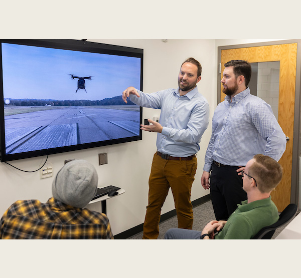 Electrum Aero's Jeffrey Bennett and Justin Jantzen stand in front of an image of a drone on a screen as they talk to two other members of their team.