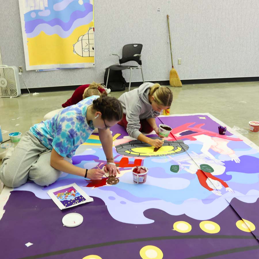 3 students paint a mural on polytab spread out on the floor 