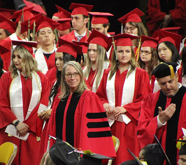 Connie Malone in Marshal regalia during a fall commencement in Millett Hall surrounded by graduating students in their graduation gowns