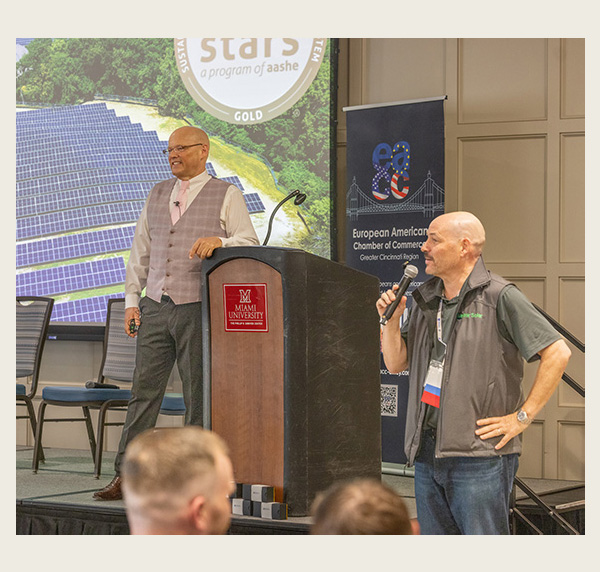 Greg Crawford near a podium in front of a large slide with a drone view of miami's solar arrays, and Fabienne Schmahl is off to the side speaking into a microphone